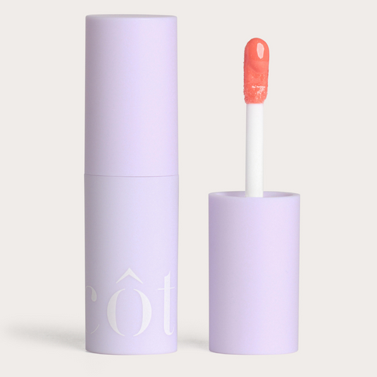 Côte Beauty The Necessity Peptide Lip Shine Branson Coral Non-Toxic Lip Gloss in a tube with an open cap beside it. 